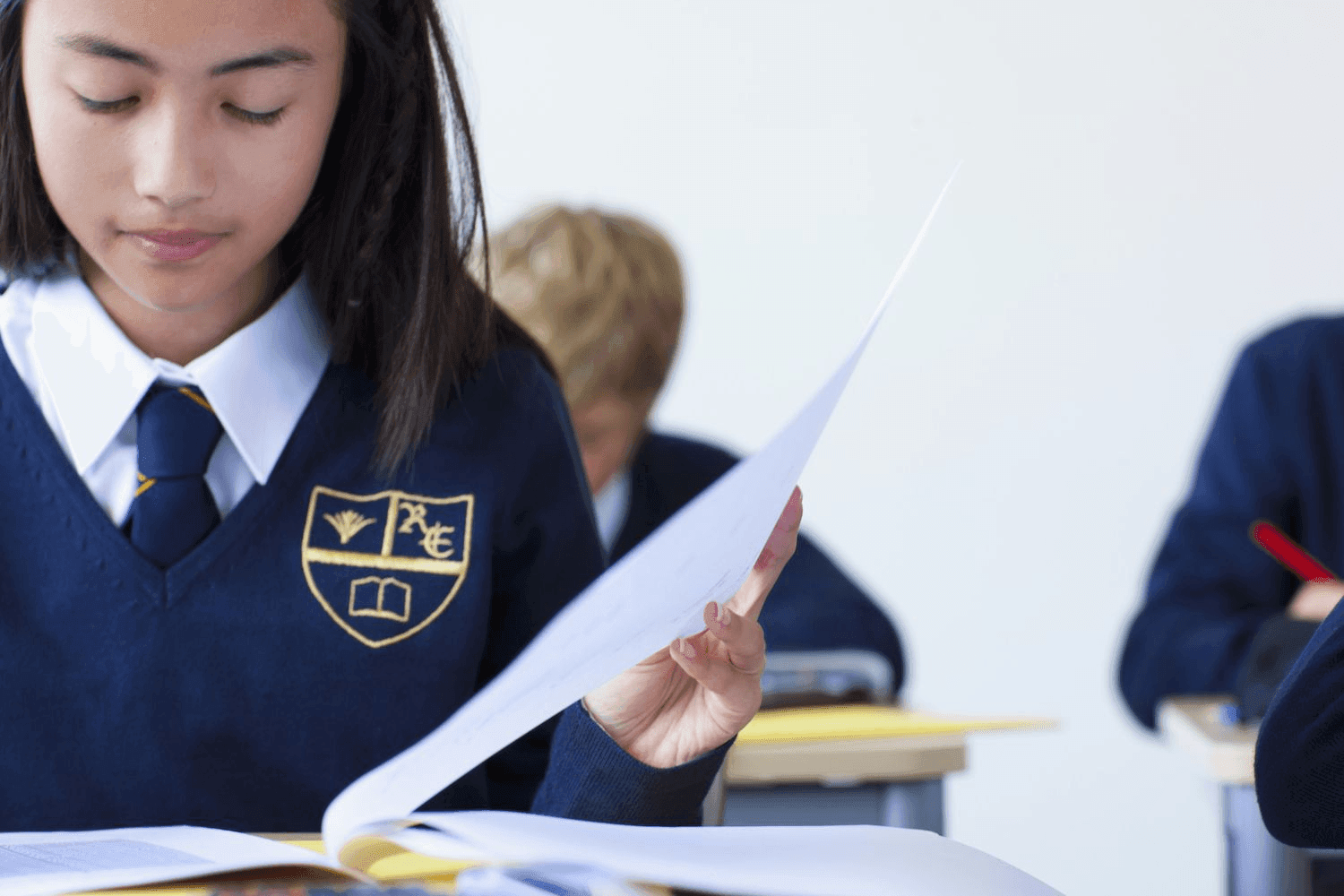 Student in a school uniform sits at a desk holding papers in a classroom setting.