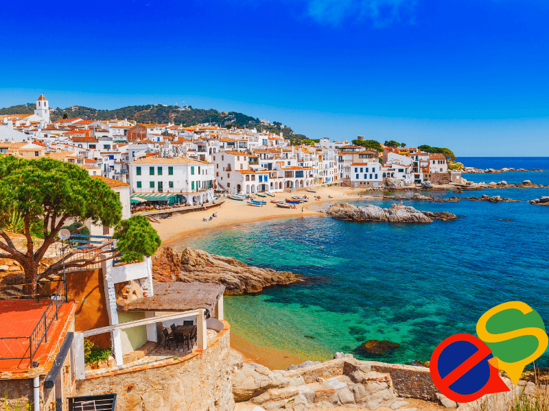 Coastal town with white buildings by a sandy beach and clear blue sea under a bright sky.