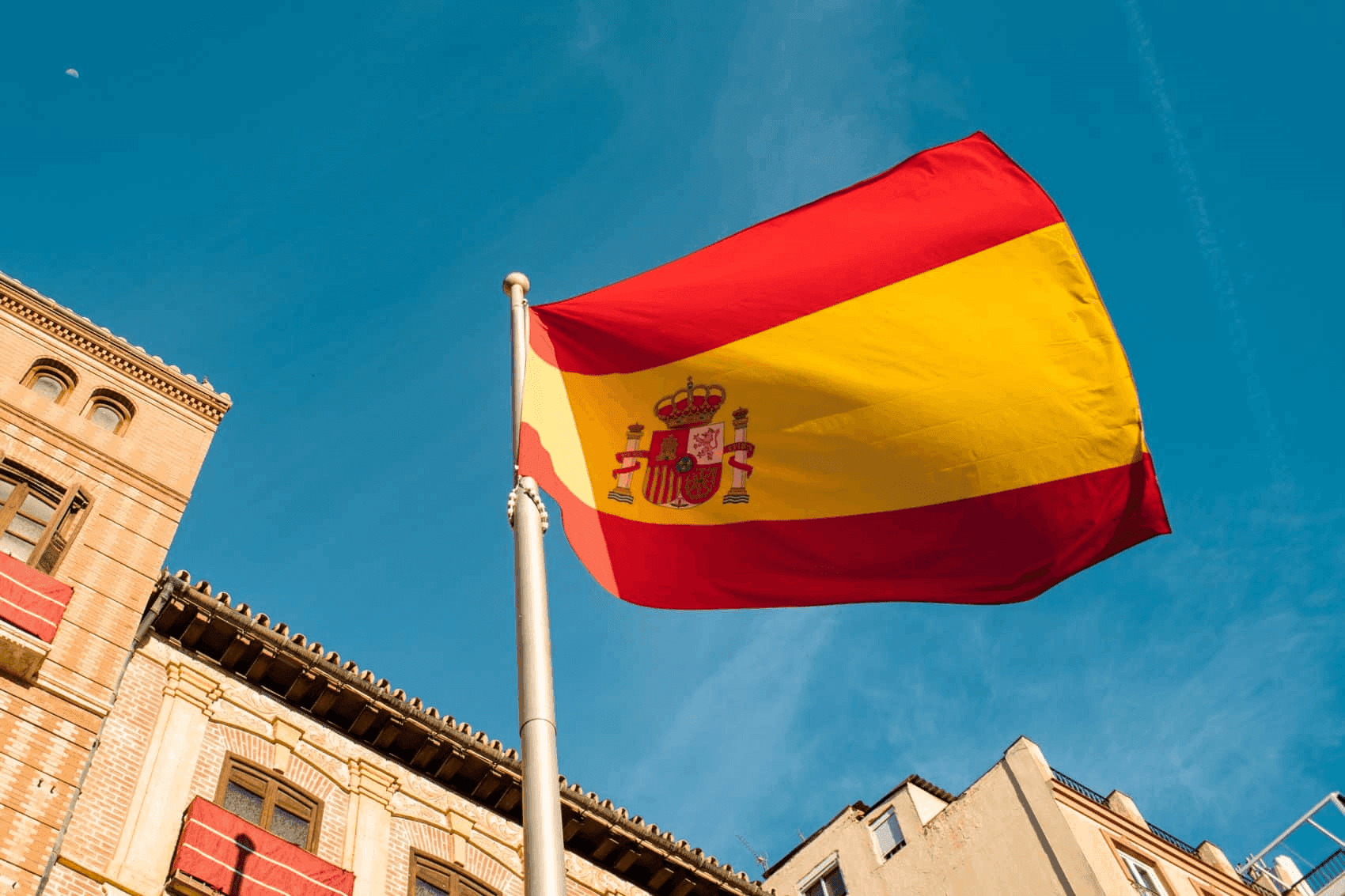 Spanish flag waving on a flagpole with buildings in the background under a clear blue sky.