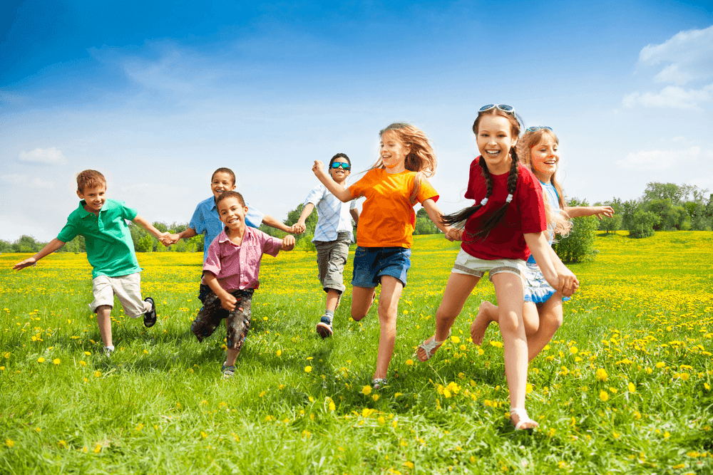 Group of joyful children running through a sunny meadow with green grass and yellow flowers.